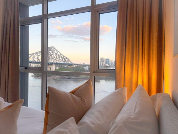 A hotel room with a large window overlooking a river and a bridge, with multiple pillows on the bed.