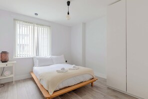 Een minimalistische slaapkamer met een houten bed, witte beddengoed, een nachtkastje met een lamp en een raam met zonwering.