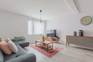 Een moderne woonkamer met een grijze bank, een houten salontafel, een rood gekleurd vloerkleed, een televisie en een minimalistische kast.