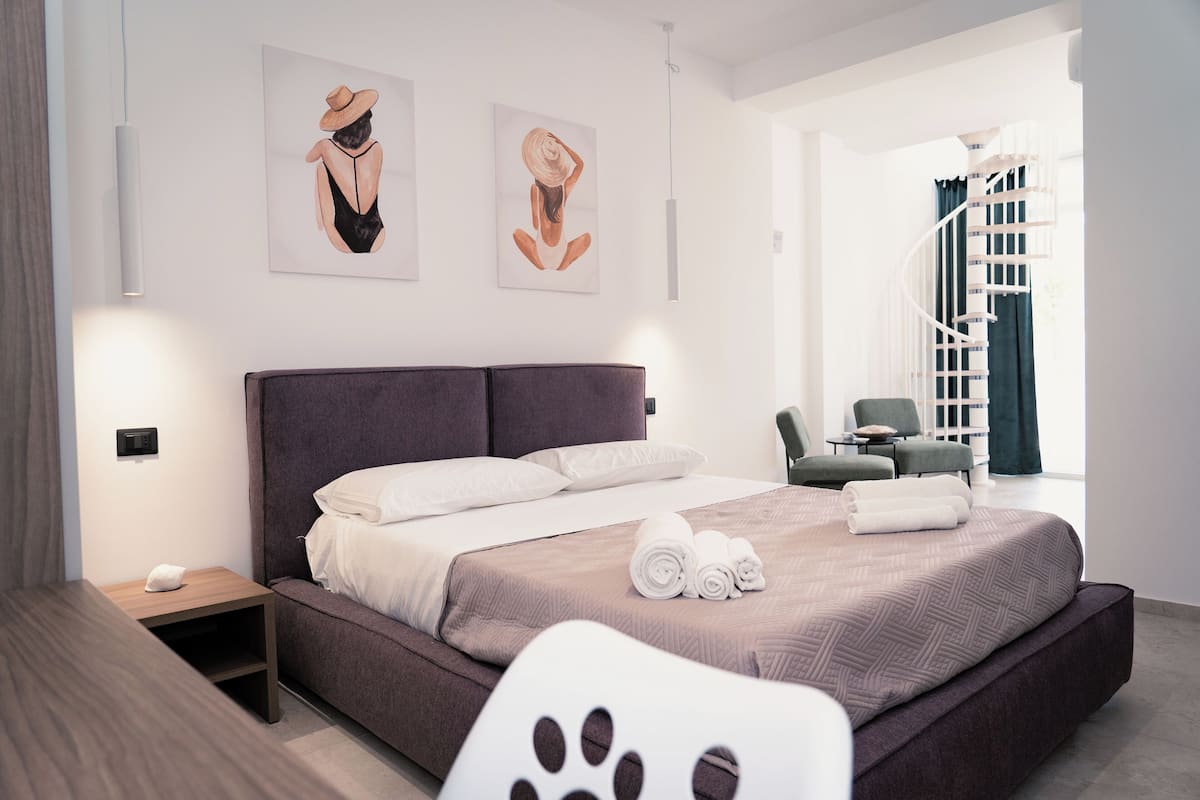 Ein modernes Hotelzimmer mit einem großen Bett, einem kleinen Nachttisch, einem Sessel und einer Treppe im Hintergrund.