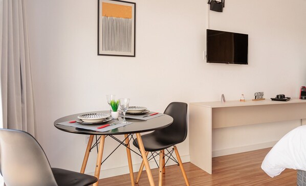 Una habitación de hotel moderna con una mesa de comedor redonda, dos sillas negras, una mesita auxiliar con una planta, un televisor de pantalla plana montado en la pared y una cama con edredón blanco.