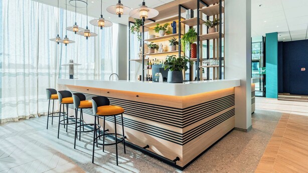 A modern bar area with a central island, bar stools, pendant lighting, and shelves with plants and bottles.