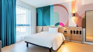 A modern hotel room with a large bed, a wooden headboard, a wall-mounted TV, and a large window with curtains.