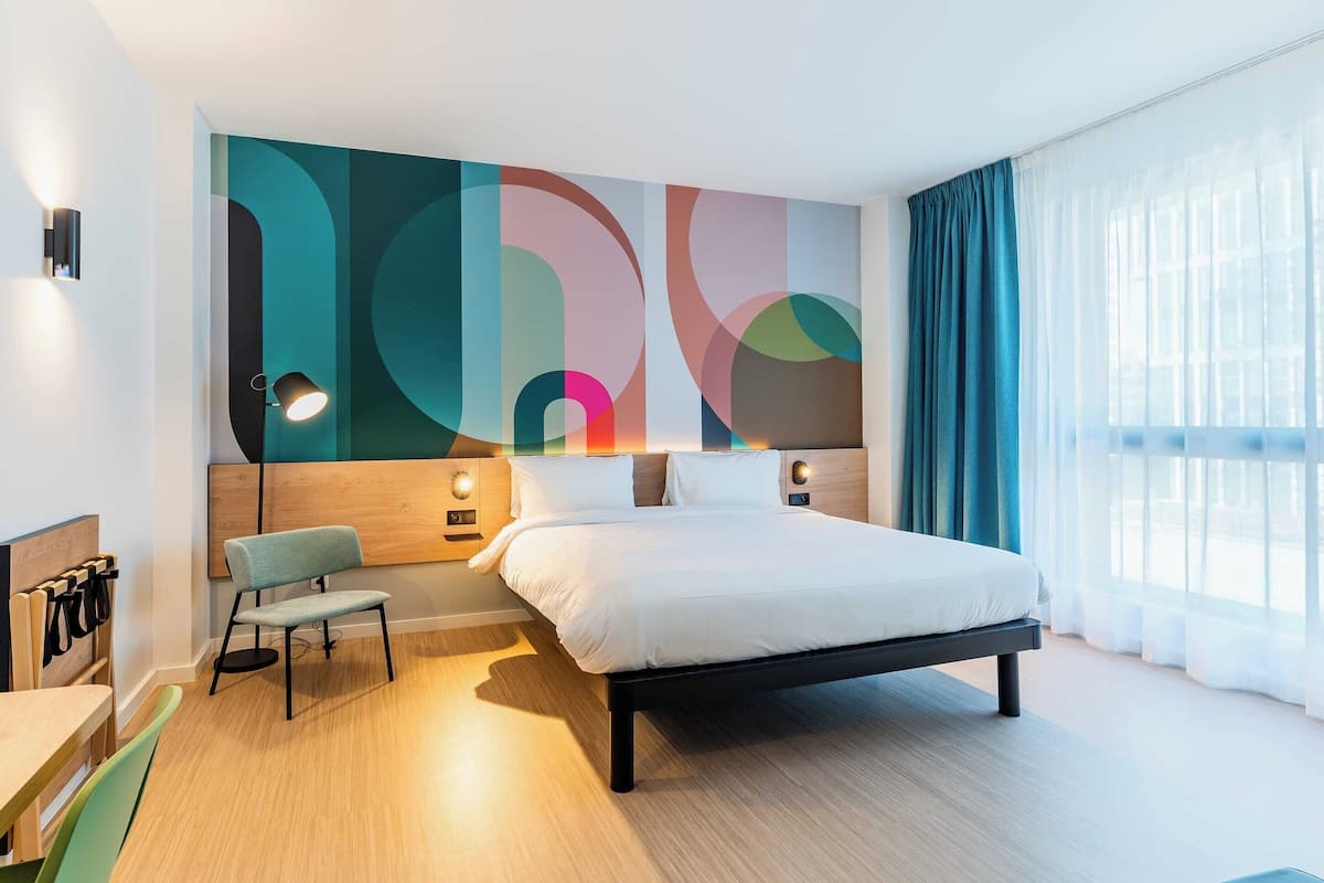 Et moderne hotelværelse med en stor seng, et siddeområde med en bænk, en vægmonteret lampe og udsigt til det frie landskab gennem lette gardiner.
