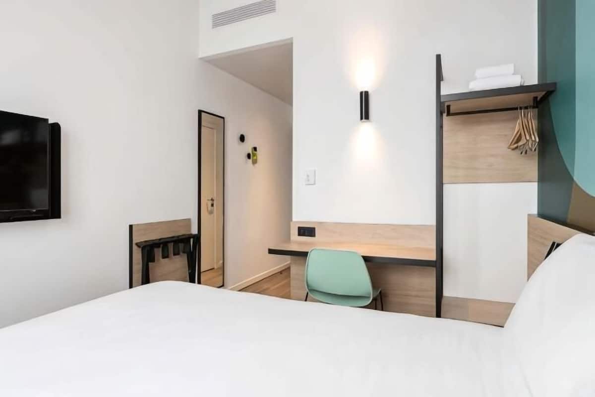 Et hotelværelse med seng, skrivebord, stol, fjernsyn og vægmonteret lampe.
