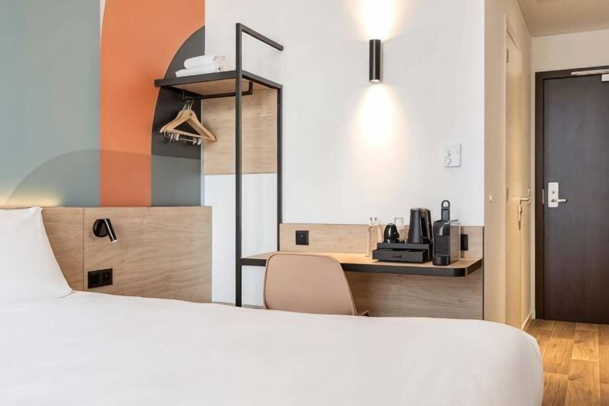 Et hotelværelse med en seng, et skrivebord med stol, et knagered og en dør.