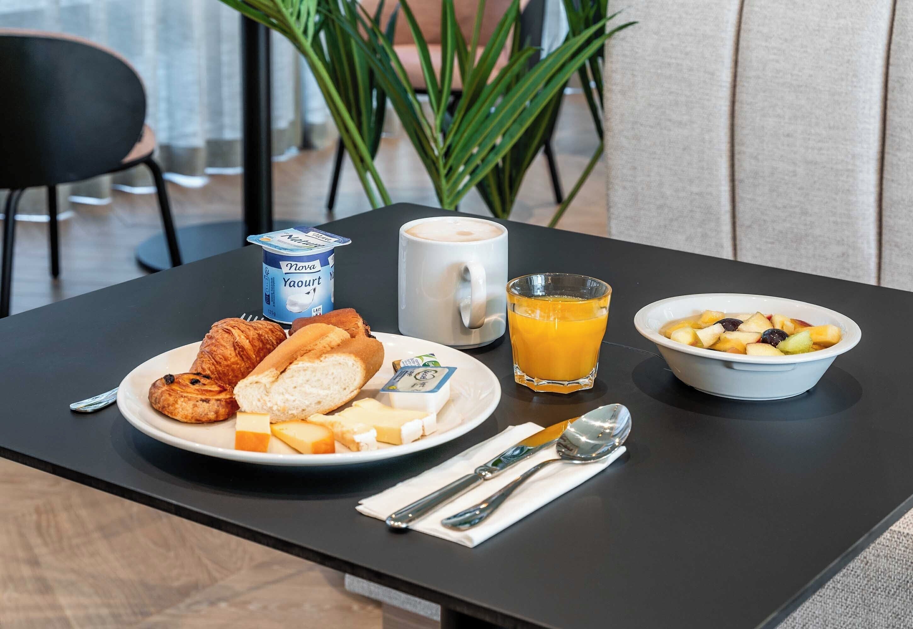A breakfast table with croissants, cheese, a coffee mug, a glass of orange juice, and a bowl of fruit.