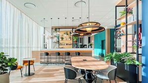 A modern hotel lobby with a wooden table and chairs, a bar area, and potted plants.