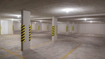 Un parking avec des piliers en béton et des bandes jaunes, un sol en béton avec des marquages au sol indiquant les places de stationnement, et une porte blanche.