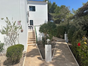 A white building with a staircase leading to a door, surrounded by greenery and a pathway. - Apartment 'Addaia' with Mountain Views, Shared Pool and Wi-Fi (Port d'Addaia)
