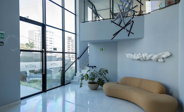 Un lobby moderno con entrada de vidrio, una escalera, una planta en maceta y un sofá color crema.