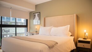 Un lit bien fait, recouvert d’une couverture beige, une table de chevet avec une lampe et une fenêtre avec des stores.