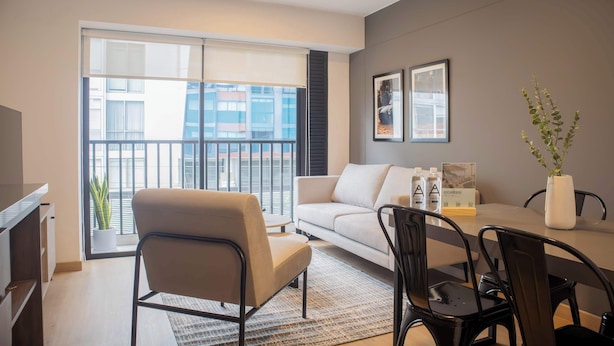A modern living room with a sofa, a dining table with chairs, a TV, and a large window with blinds. - w Striking 1BR Duplex in Miraflores (Lima)