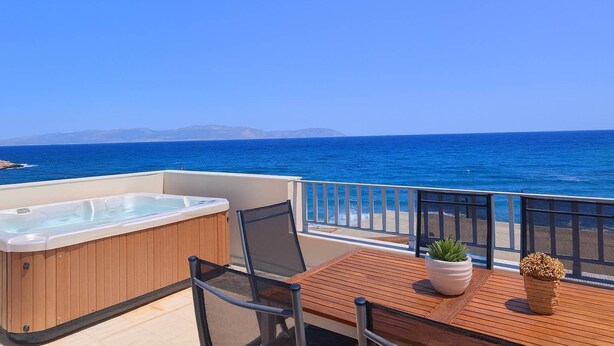 A balcony with a hot tub, wooden table, chairs, and potted plants, overlooking a clear blue sea and distant mountains. - Beachfront Ela Luxury Stay With Jacuzzi Views (Kithira)
