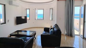 A living room with a flat-screen TV, a leather sofa, a glass coffee table, and a view of the sea through open windows. - Beachfront Ela Luxury Stay With Jacuzzi Views (Kithira)