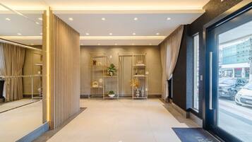 A modern hotel lobby with a reception desk, a glass door leading outside, and a decorative plant arrangement.