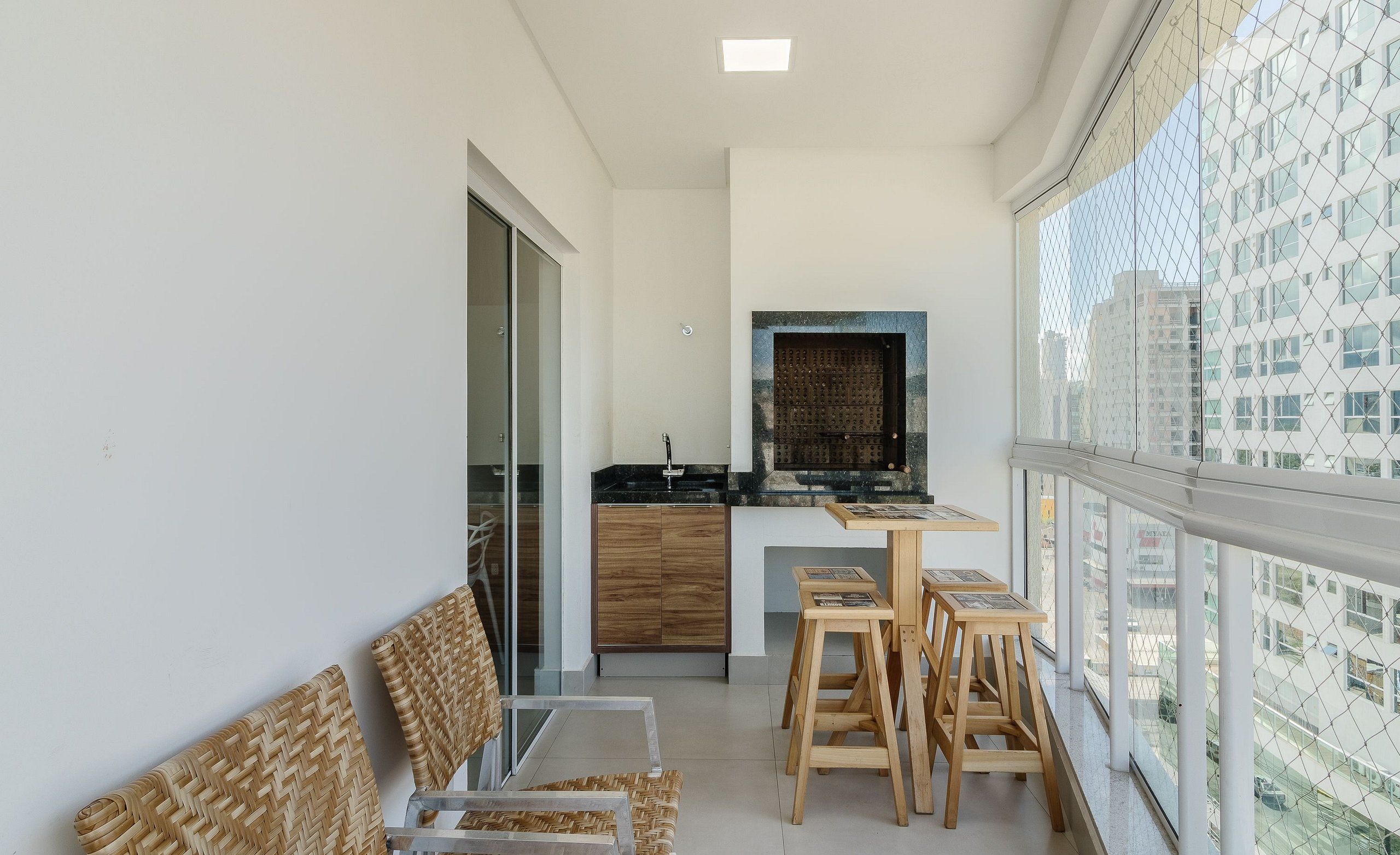 Un balcon avec des fauteuils en osier, une petite table en bois et un coin cuisine doté d’un plan de travail noir et de placards en bois.