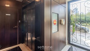 En elevator med dør i træ og glasrude, et skilt der viser udgang, samt udsigt til det fri gennem et metalgitter.