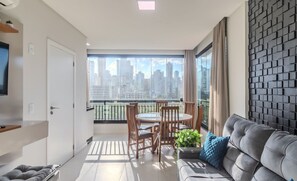 A modern living room with a grey sofa, a dining table with chairs, and a large window with a city view. - Apart With Barbecue 350m From the Beach Pdp0301 (Porto Belo)