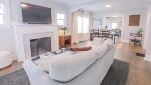 A modern living room with a fireplace, a large flat-screen TV, a sofa, and a dining area with chairs and a table.