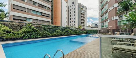 Una piscina con cubierta de madera y barandales de metal, rodeada de follaje y edificios modernos.