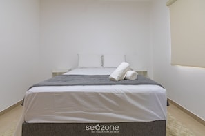Un lit bien fait, avec une couverture grise et une serviette blanche roulée, un oreiller blanc et une tête de lit blanche.