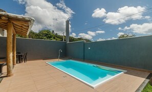 Una piscina rectangular con borde blanco, rodeada por un patio con baldosas. A la izquierda hay una estructura de pérgola de madera y, al fondo, un tanque de agua alto y cilíndrico.