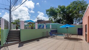 Zona de piscina con terraza de madera, una sombrilla azul y una escalera que lleva a una entrada con portón.