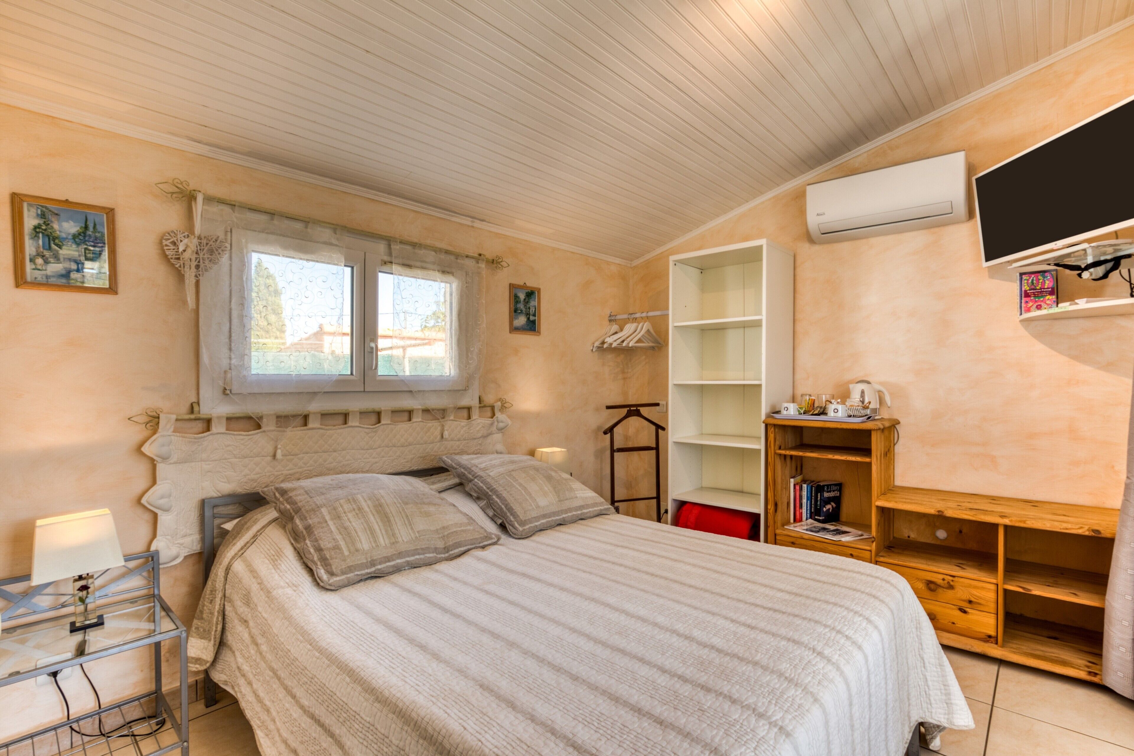 A bedroom with a bed, a bedside table, a window with curtains, a wall-mounted TV, and a shelf with books.
