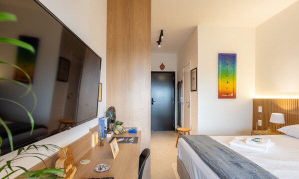 A hotel room with a bed, a desk with a computer, a large mirror, and a wall-mounted lamp.