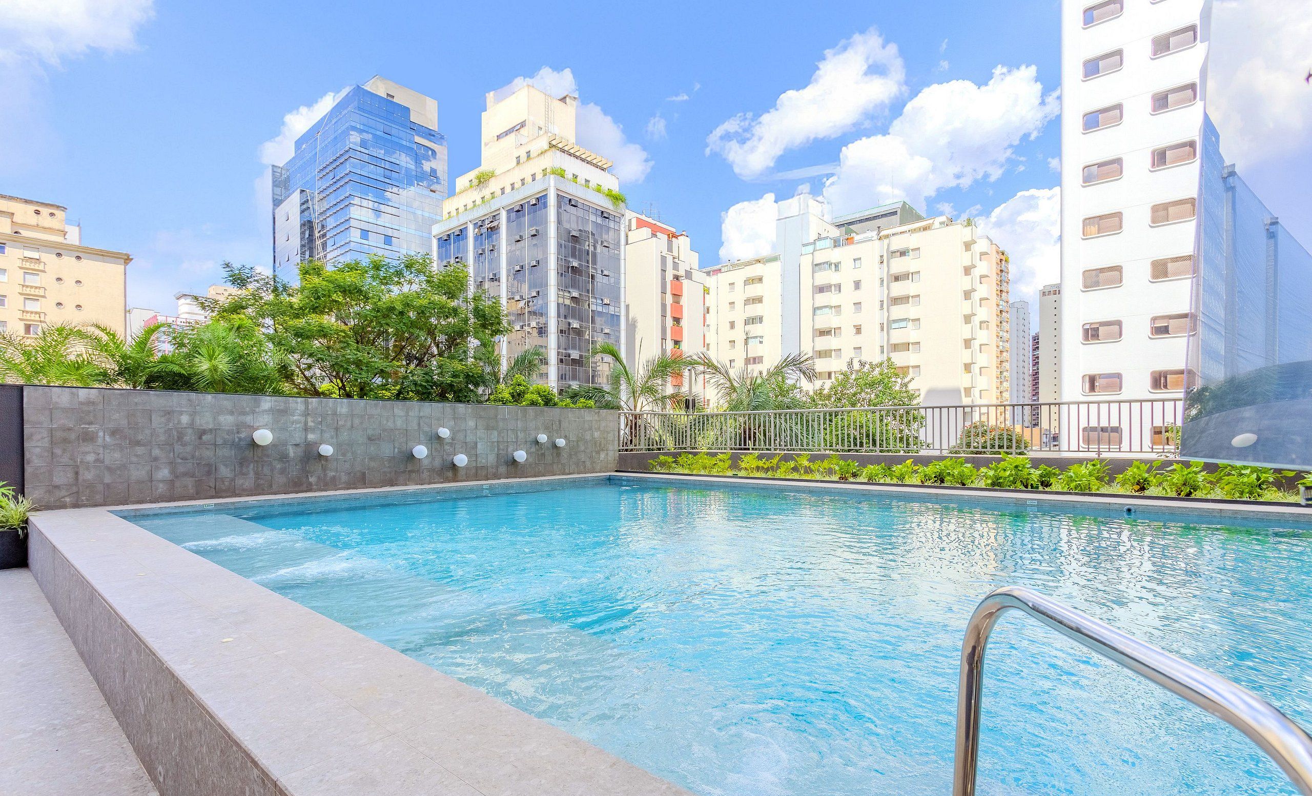 A rooftop swimming pool with a cityscape view, surrounded by greenery and modern buildings.