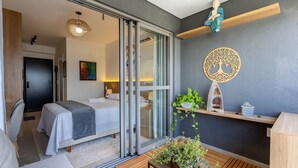 A modern hotel room with a sliding glass door leading to a balcony, a bed with a gray blanket, a small table with a plant, and a decorative wall piece.