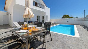 A modern outdoor dining area with a pool, white walls, and a clear blue sky.