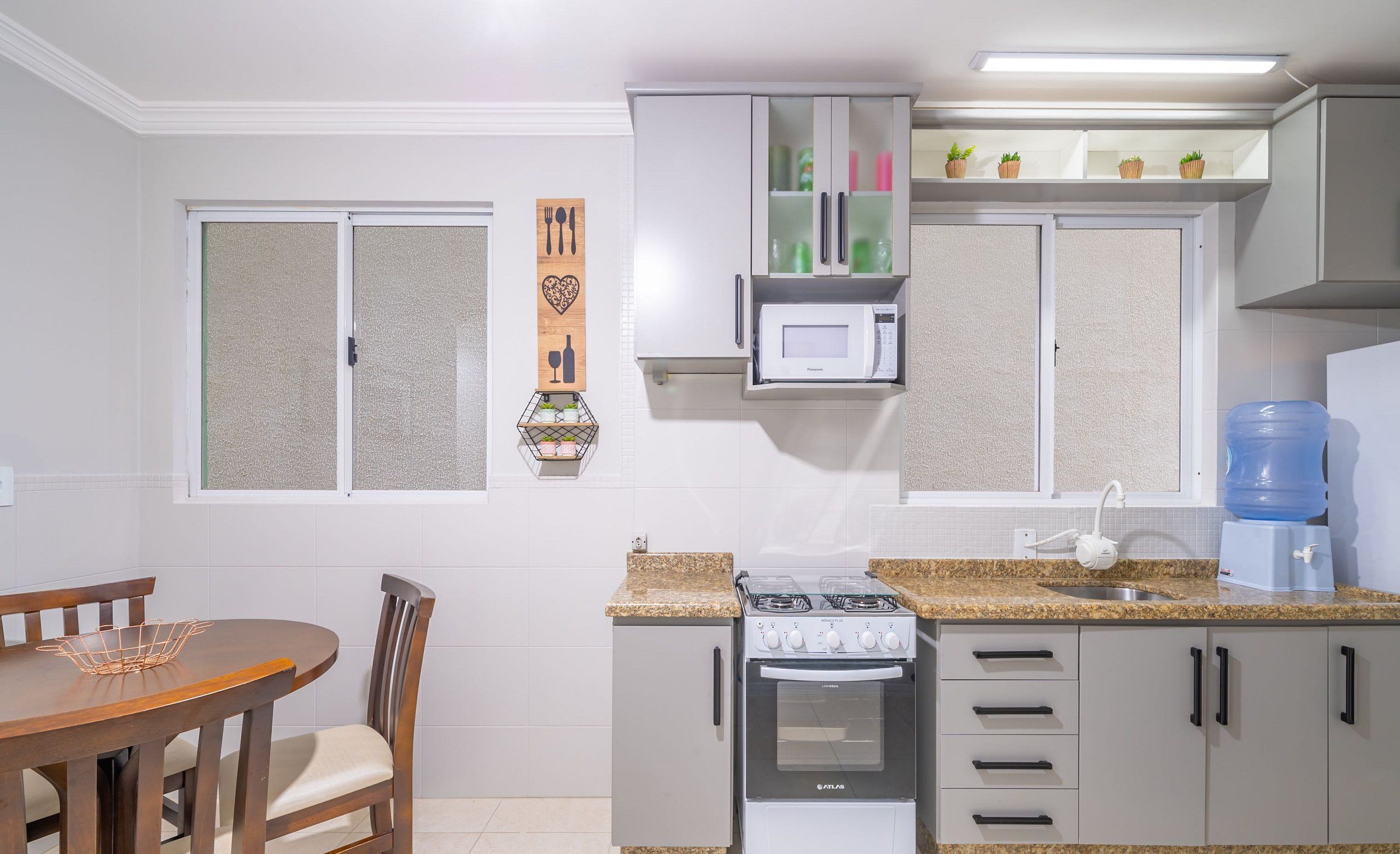 A compact kitchen with a gas stove, oven, microwave, and a water dispenser. There is a dining table with chairs and a decorative wall hanging.