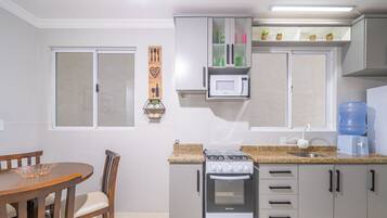 A compact kitchen with a gas stove, oven, microwave, and a water dispenser. There is a dining table with chairs and a decorative wall hanging.