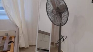 A room with a standing fan, a wooden bed, a white door, and a window with curtains.