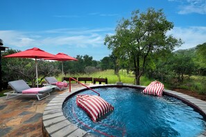 A swimming pool with lounge chairs and red umbrellas, surrounded by trees and a stone deck. - Dzombo Camp (Pilanesberg National Park)