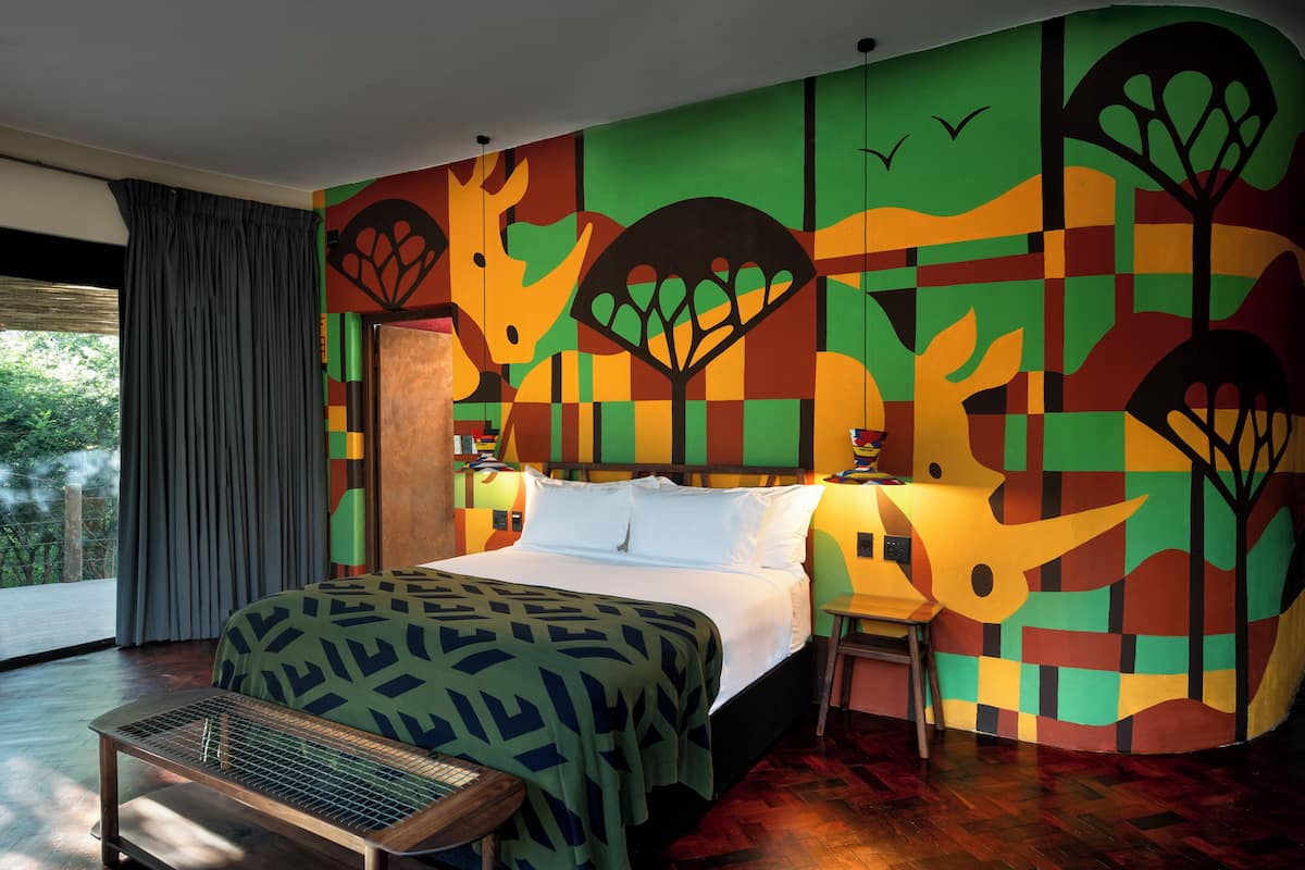 Habitación de hotel con una cama grande, una mesita de noche de madera, una ventana con cortinas y un mural abstracto y colorido en la pared.