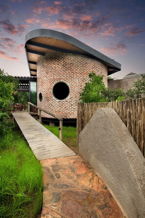 A modern house with a brick exterior, circular windows, and a wooden deck leading to the entrance. - Dzombo Camp (Pilanesberg National Park)