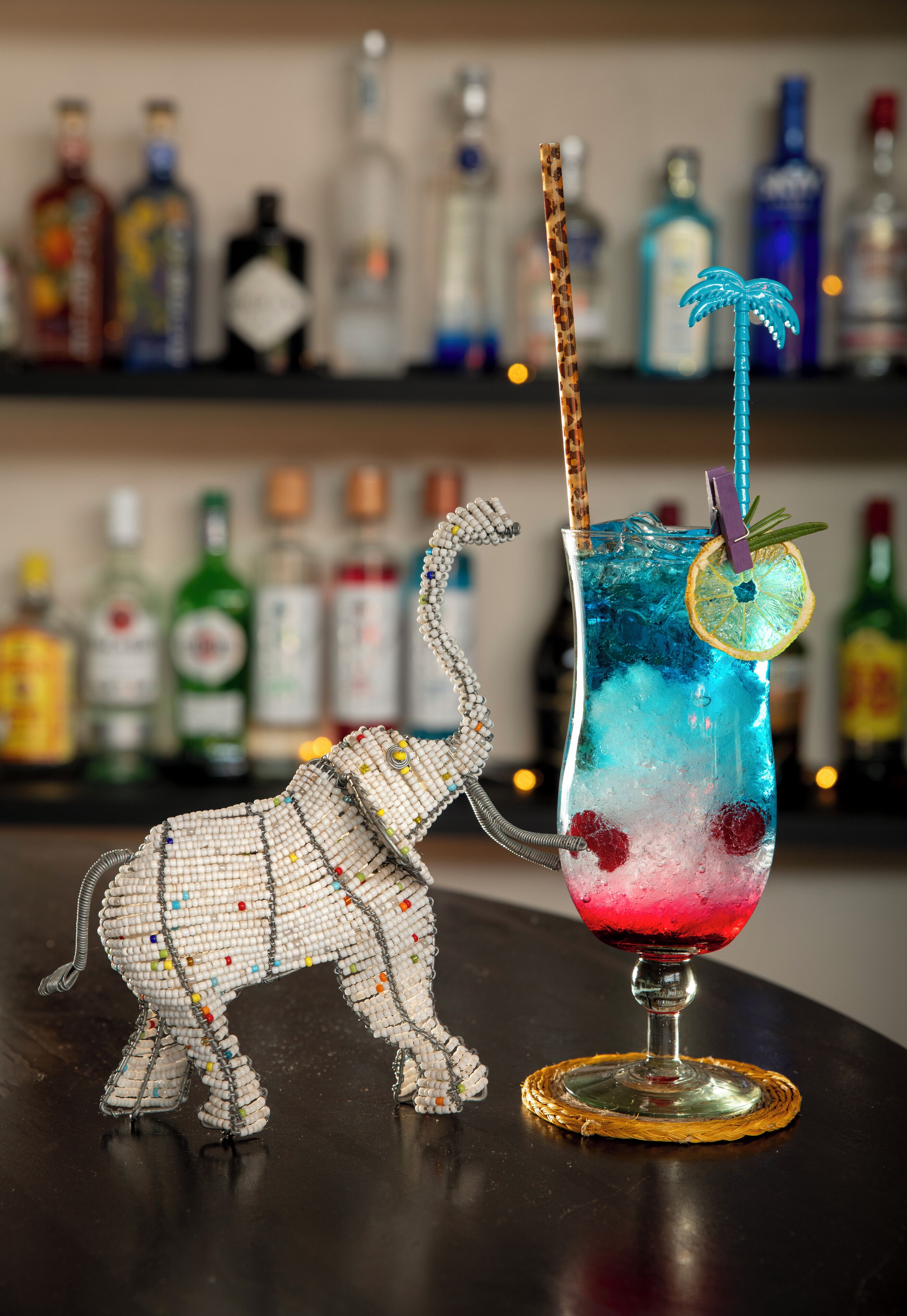 A cocktail with a straw and a decorative umbrella, a beaded elephant figurine, and a bar with various bottles in the background.