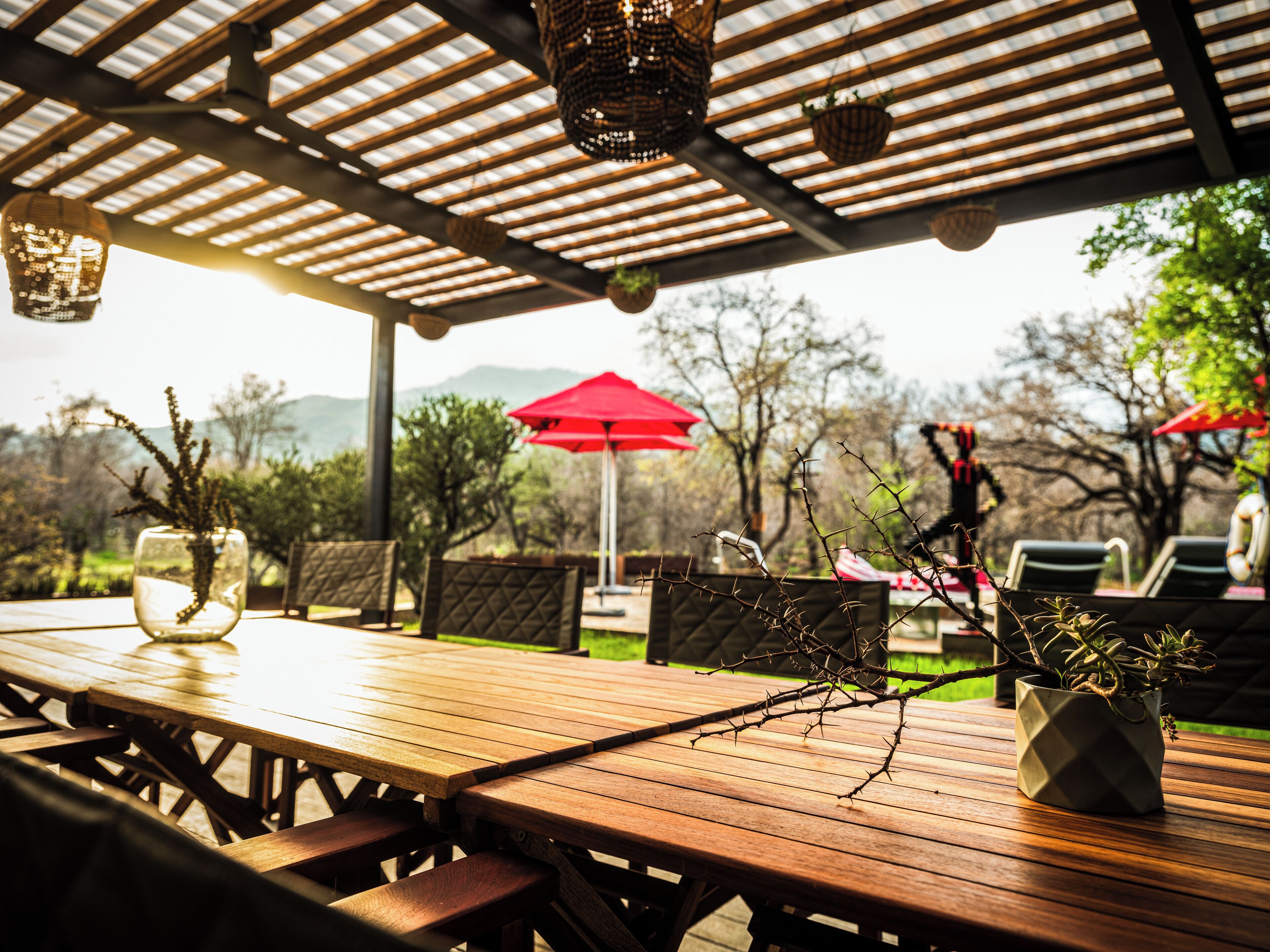 An outdoor patio with wooden tables and chairs, a red umbrella, and hanging lanterns.