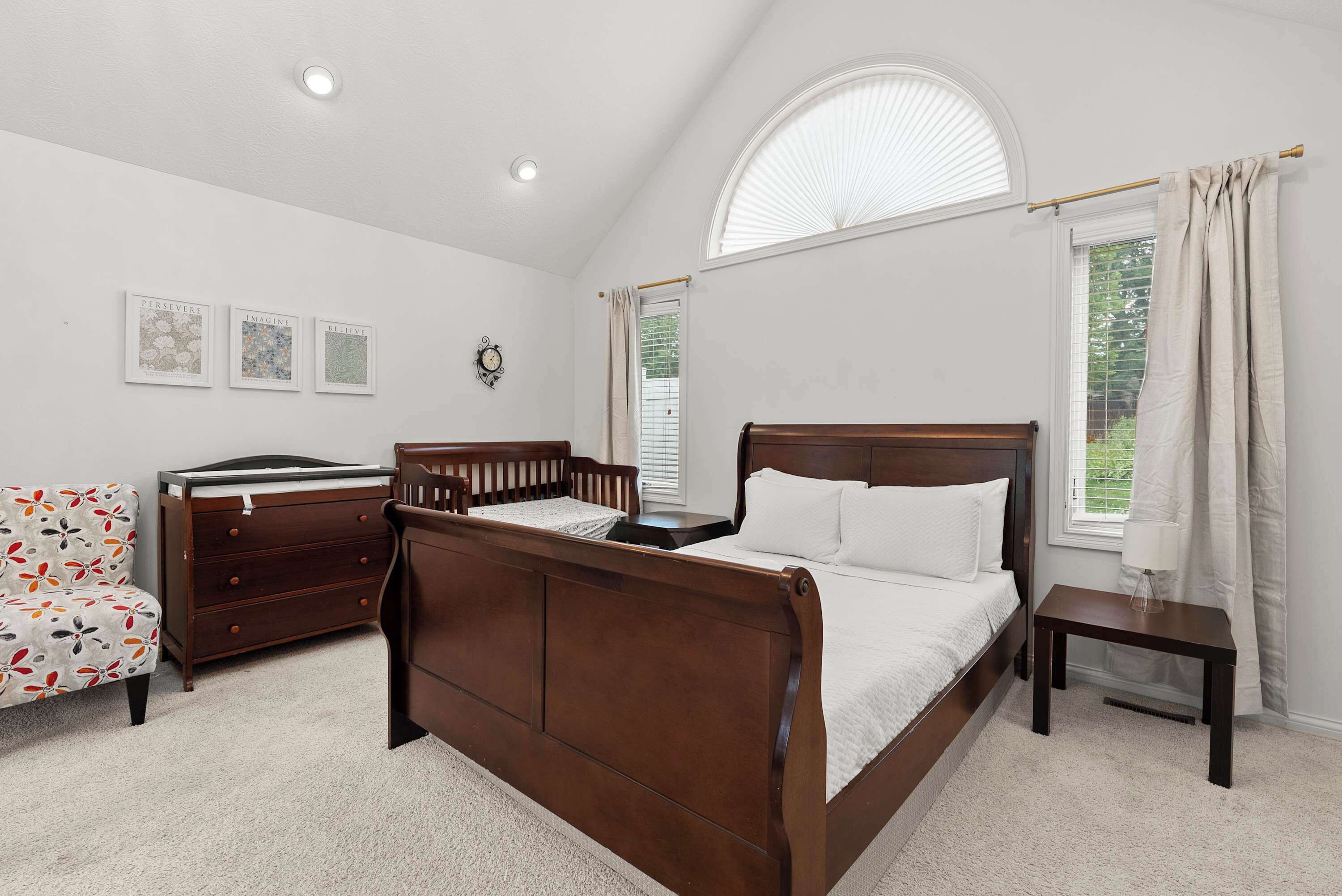 A bedroom with a wooden bed, a dresser, a chair, and a small table. There are three framed pictures on the wall and a window with curtains.