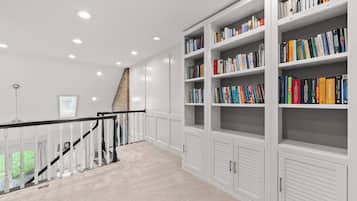Une bibliothèque spacieuse, avec des étagères blanches remplies de livres, un escalier doté d’une rampe en verre et un mur de briques en arrière-plan.