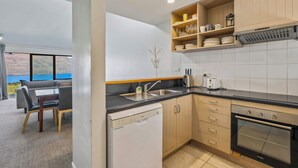 A modern kitchen with wooden cabinets, a dishwasher, a stove, and a dining area with a view of the outdoors. - Outlook Oasis, Uninterrupted Lake Views (Queenstown)