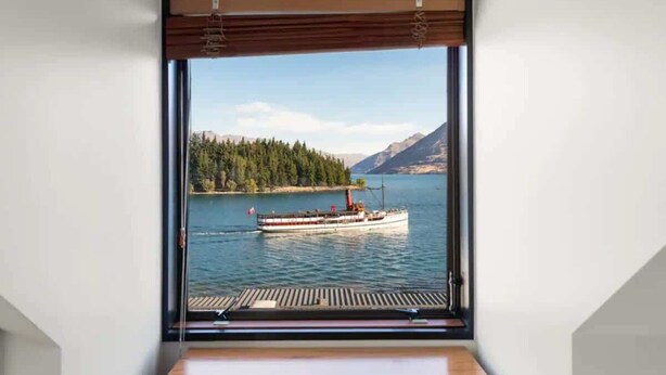 Una habitación con un ventanal grande que da a un lago, un barco y colinas boscosas.