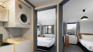 Una habitación de hotel con lavadora, una cama, un escritorio con silla y un ventanal con cortinas.