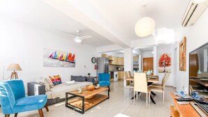 A modern living room with a sofa, blue armchair, and coffee table. There is a dining area with a table and chairs. A kitchen is visible in the background. - Nissi Apartment 102 (Ayia Napa)
