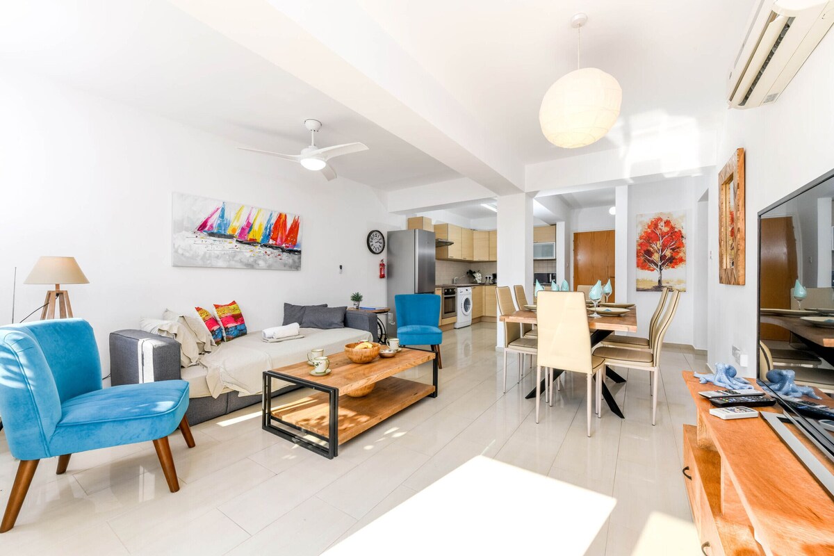 Una sala de estar moderna con un sofá, un sillón azul y una mesa de centro. Se observa un comedor con una mesa y sillas. Se ve una cocina al fondo.