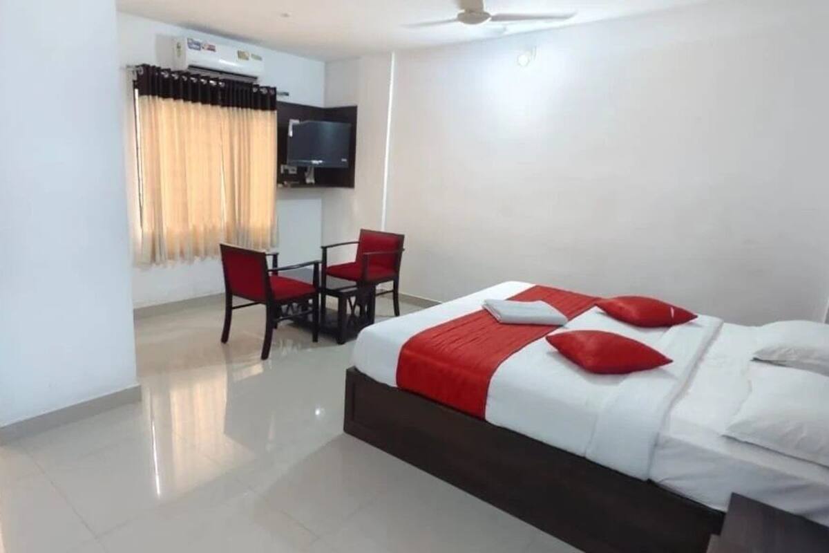 Quarto de hotel com uma cama, almofadas vermelhas, uma televisão e um espaço de estar com cadeiras.