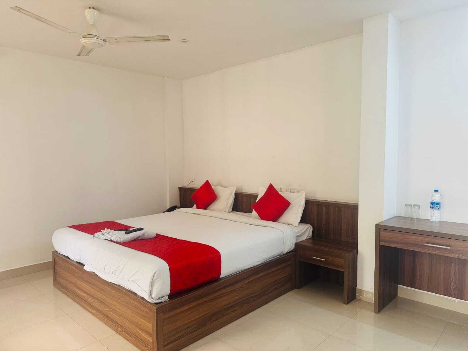 Une chambre d’hôtel avec un lit en bois, du linge de lit blanc, des oreillers rouges, une table de chevet, un bureau et un ventilateur de plafond.